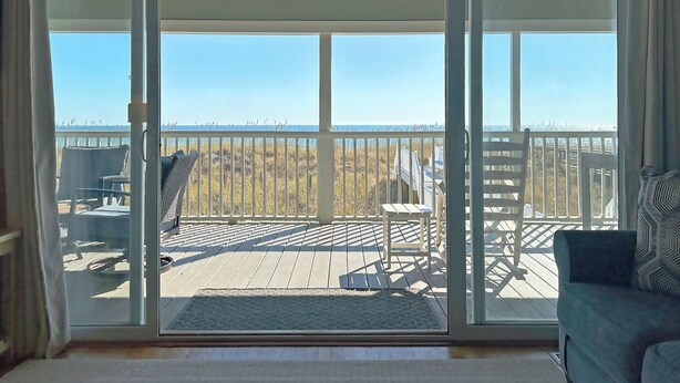 Habitació amb porta corredissa de vidre que dona a una terrassa amb mobles d'exterior i vista a una platja.
