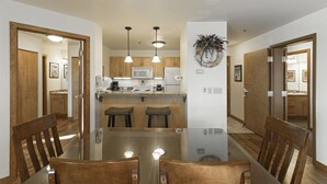 A dining area with a glass table, wooden chairs, pendant lights, and a kitchen in the background. - Some of the best direct ski access on the mountain!! 2 bedroom with beautiful valley views!! (Whitefish)