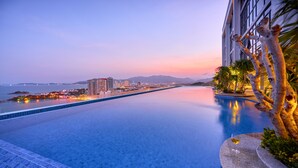 An infinity pool with a view of a cityscape and mountains at dusk. - Scenia Bay Residences (Bac Nha Trang)
