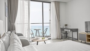 A modern hotel room with a large bed, a desk, a chair, a small table with a glass of water and a book, and a TV on a wooden stand. - Scenia Bay Residences (Bac Nha Trang)