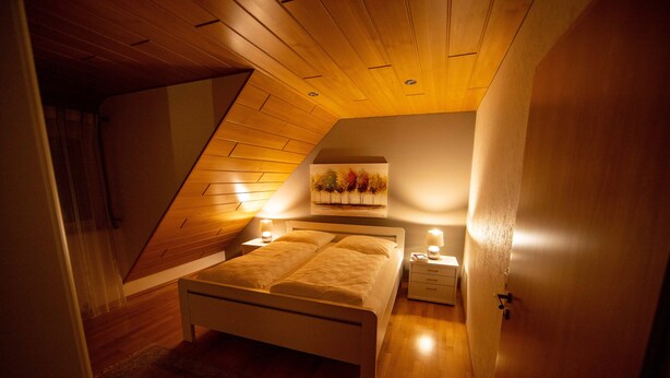 A bedroom with a sloped ceiling, a bed with white bedding, two bedside tables with lamps, and a wall-mounted artwork of trees. - FeWo Studenik (Neuried)