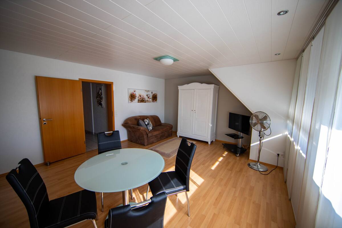 A room with a round table, four chairs, a sofa, a cabinet, a TV, and a fan.