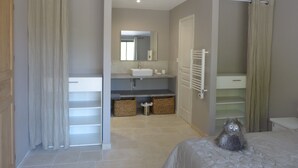 A modern bedroom with a bed, a nightstand, a dresser, a mirror, and a bathroom with a sink and towel rack.