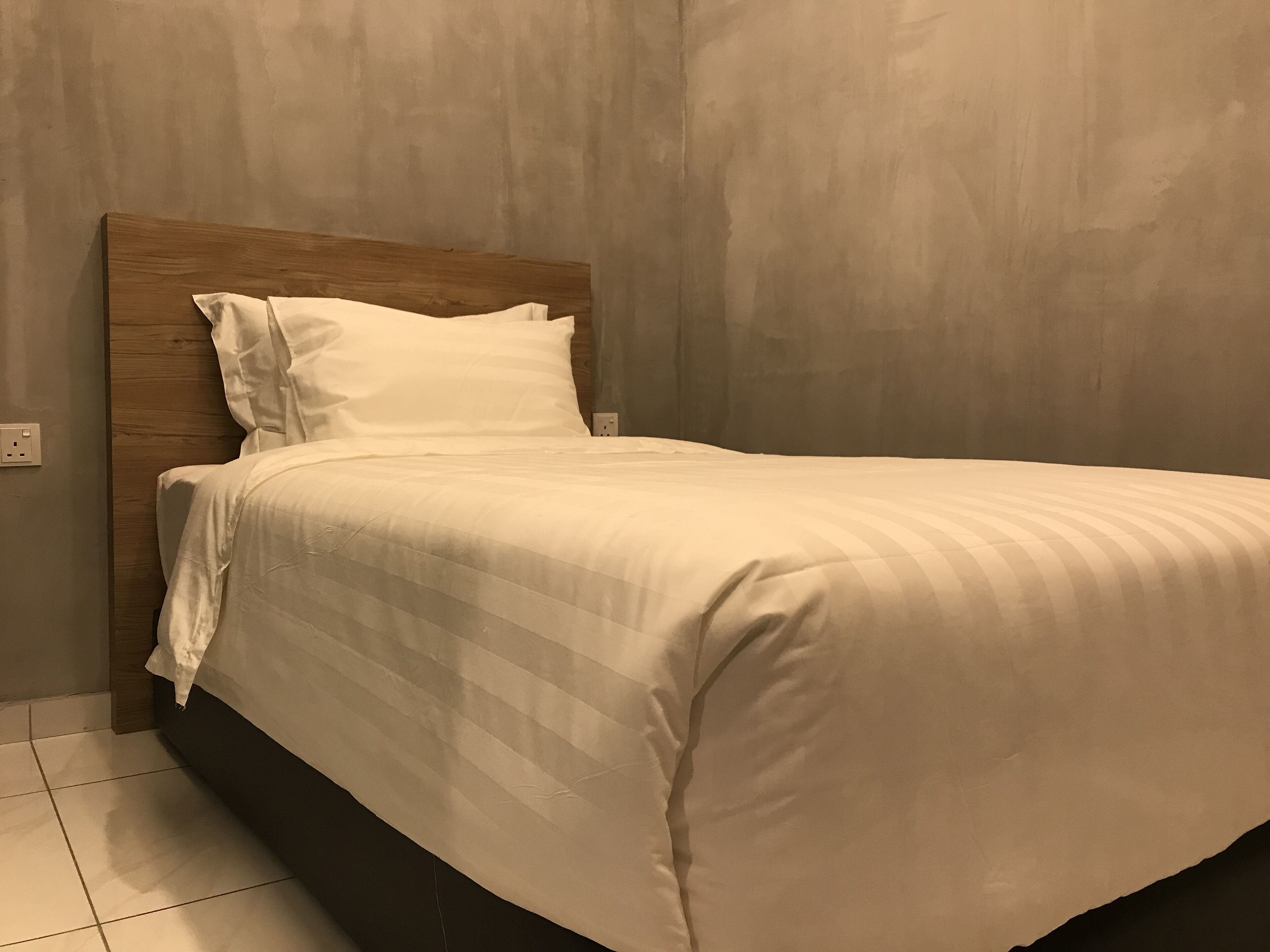Una cama bien tendida con cabecera de madera en una habitación con paredes lisas y suelo de baldosas.