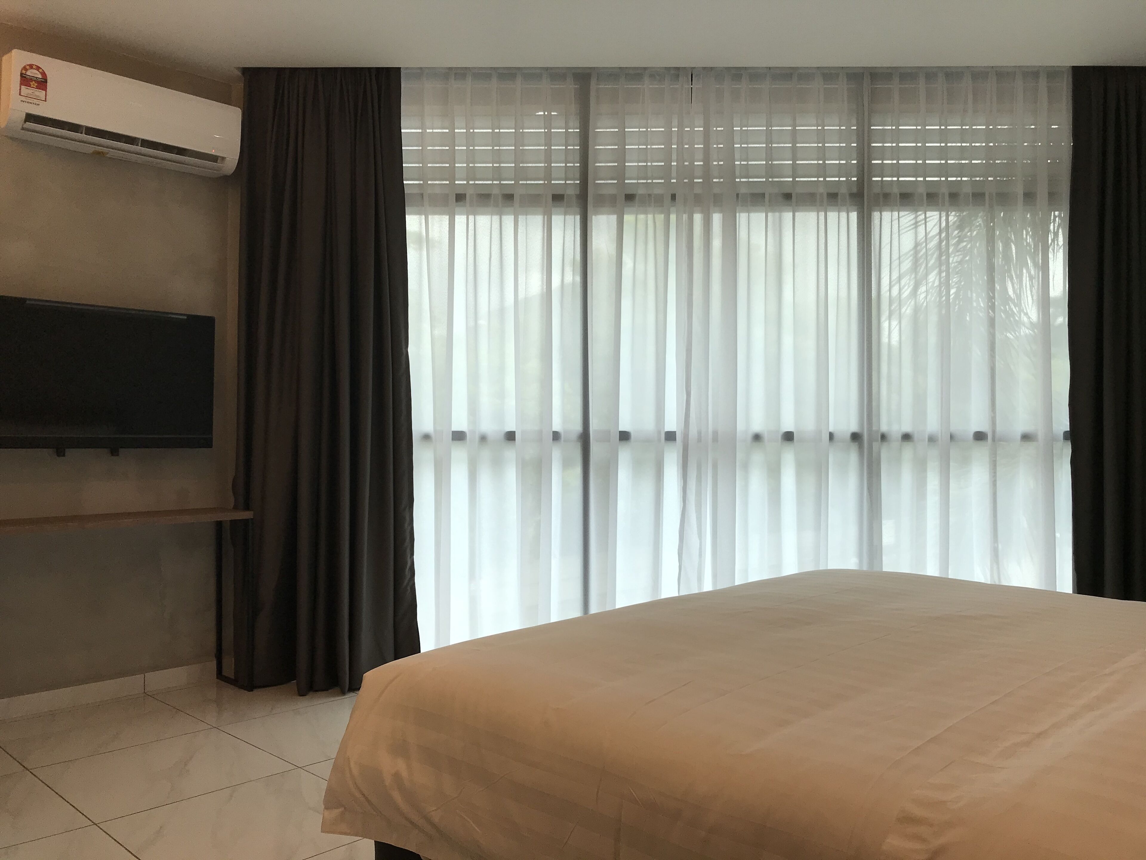 A hotel room with a bed, a flat-screen TV mounted on the wall, and a large window with sheer curtains.