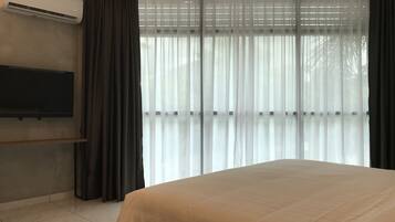 A hotel room with a bed, a flat-screen TV mounted on the wall, and a large window with sheer curtains.
