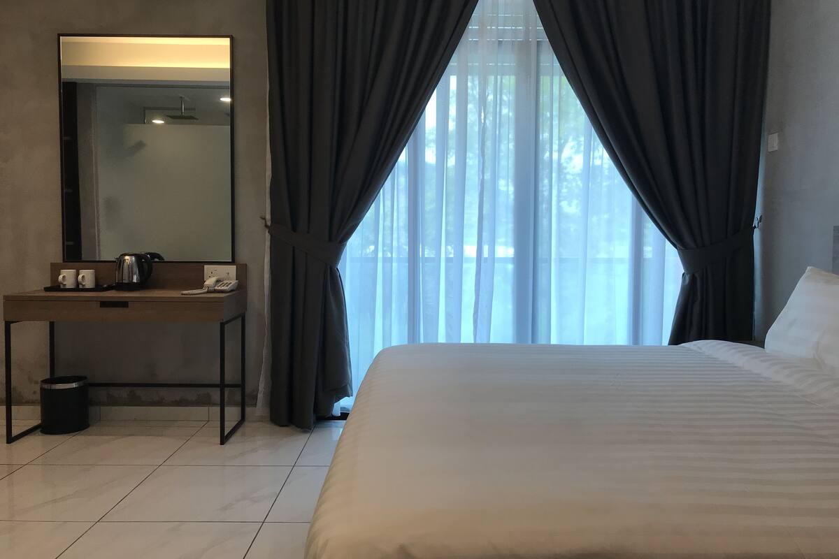 Habitación de hotel con cama, mesita de noche con teléfono y hervidor, espejo y ventana con cortinas.