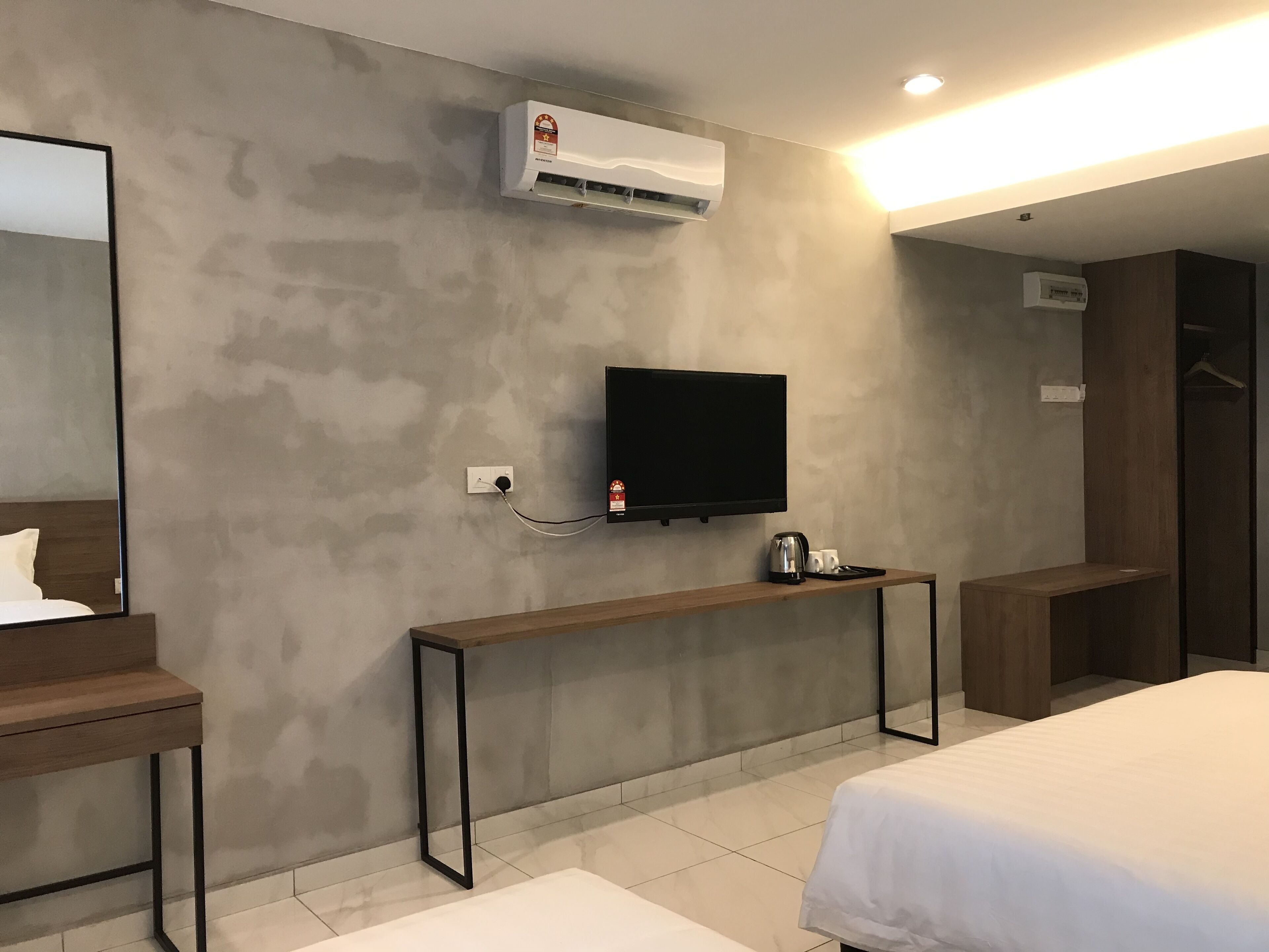 Habitación de hotel con una cama grande, una mesita de noche con tetera, un televisor de pantalla plana montado en la pared y un equipo de aire acondicionado.