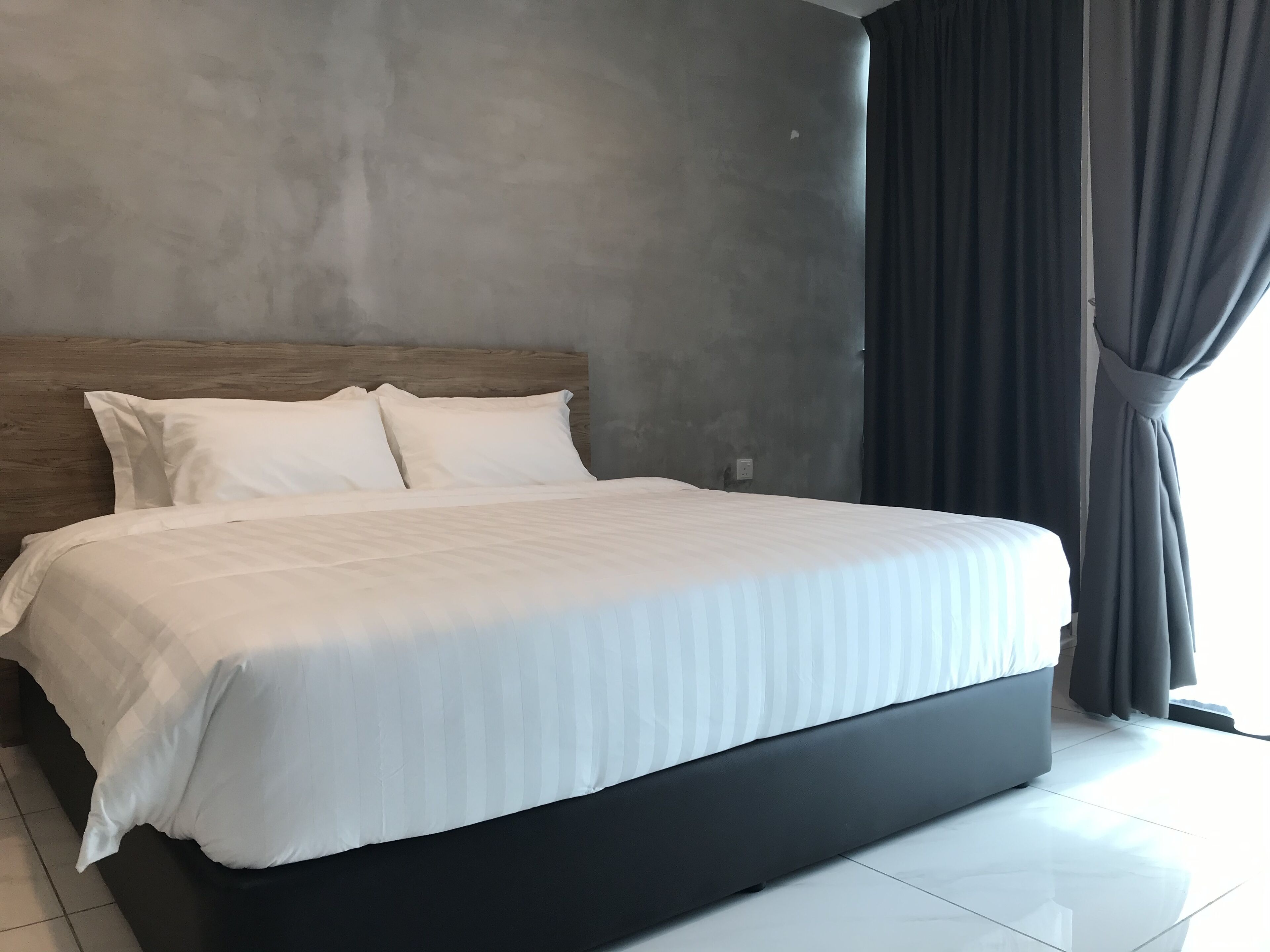 Una cama bien tendida con sábanas blancas en una habitación de hotel moderna, con una pared de concreto gris y una cortina oscura en la ventana.