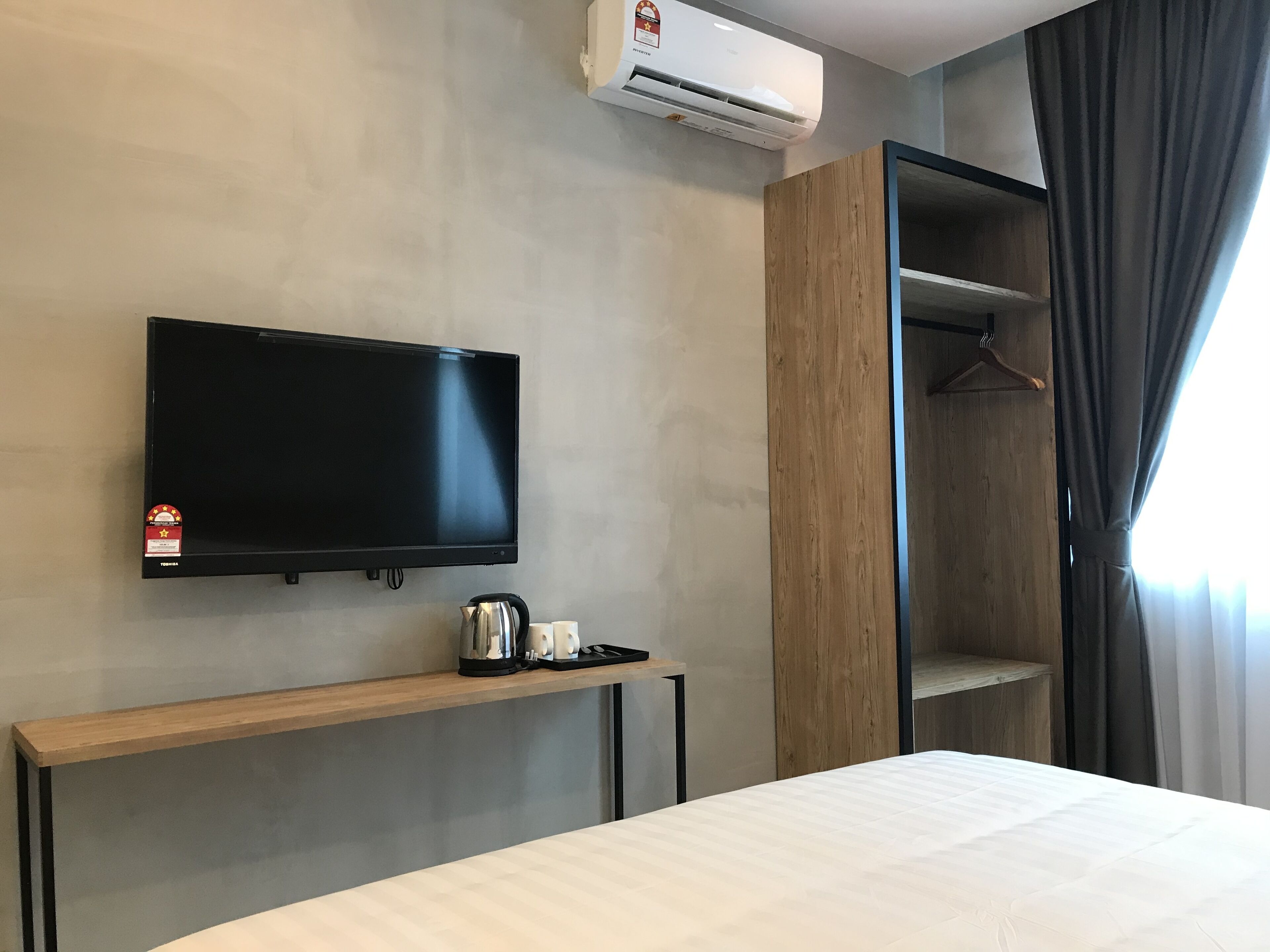 Camera d'albergo con televisore a schermo piatto montato a parete, un ripiano in legno con bollitore e tazze, un letto con materasso bianco e un armadio con appendiabiti.