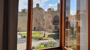 Vista per una finestra d'un edifici històric, amb un gran arbre al primer pla, un pati pavimentat i un cel clar.
