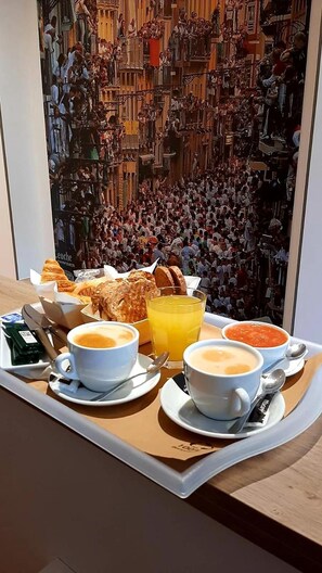 A breakfast tray with croissants, soup, and juice on a table with a view of a crowded square.
