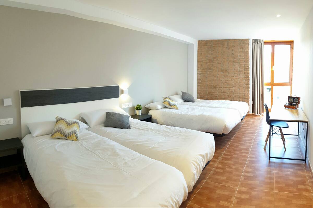 Quarto de hotel com duas camas, uma escrivaninha com cadeira, parede de tijolos e janela com cortinas.