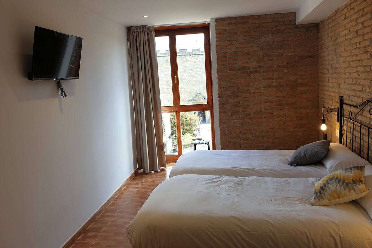 Quarto com uma grande janela, televisão de tela plana na parede, cama com travesseiros e mesa de cabeceira com abajur.