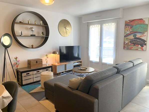 A modern living room with a grey sofa, a flat-screen TV, a round mirror, and a bookshelf.