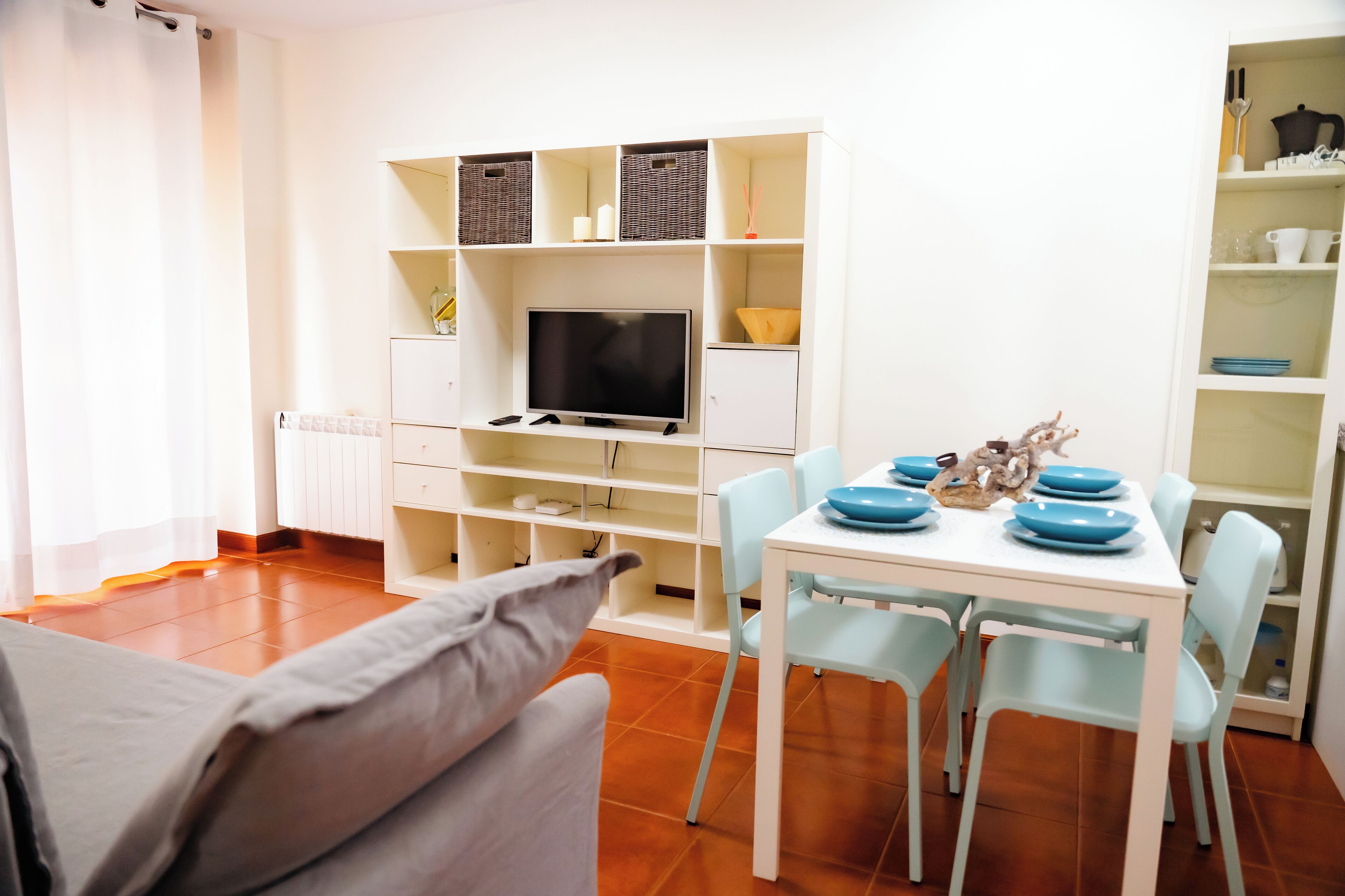 A modern living room with a TV, white shelves, a dining table with blue plates, and a sofa.