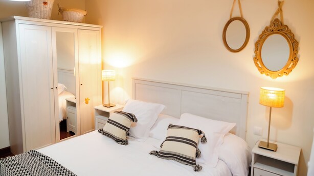 A neatly made bed with white linens and striped pillows, a white wardrobe, and two bedside lamps. - Apartamentos Costa Norte (San Vicente de la Barquera)