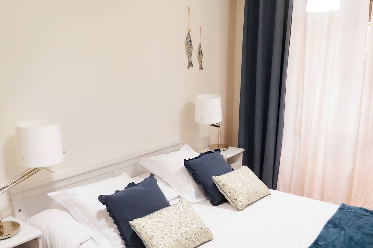Un lit bien fait, avec du linge de lit blanc et des coussins décoratifs, une table de chevet avec une lampe et une fenêtre avec des rideaux.