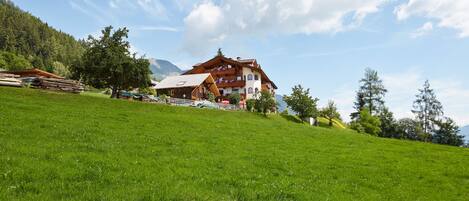 A large wooden chalet with multiple balconies, set on a grassy hillside with mountains in the background.