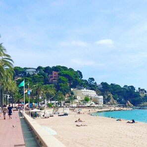 A coastal beach scene with a paved walkway, beach umbrellas, and people relaxing on the sand.