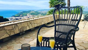 A table with a glass of orange juice and a bowl, a chair, and a view of the sea and town. - Panoramic Sea View and Private Pool (Lloret de Mar)