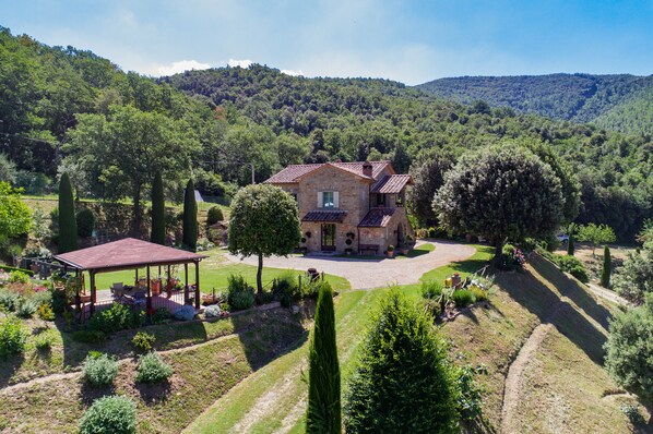 Une maison en pierre avec un toit en tuiles, entourée d’un jardin bien entretenu et offrant une vue dégagée sur une colline boisée.