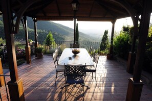 Une terrasse extérieure couverte, dotée d’une table et de chaises, entourée de plantes en pot et offrant une vue panoramique sur les collines.
