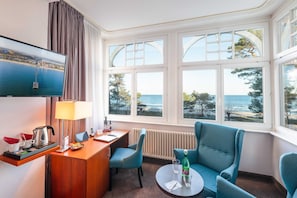 A hotel room with a large window, a desk with a lamp, a television, and two blue armchairs. - Double room - Hotel Villa Belvedere (Binz)