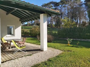 Eine Terrasse mit Tisch und Stühlen, ein überdachter Bereich mit einem Fenster und Blick auf eine Rasenfläche mit Bäumen.