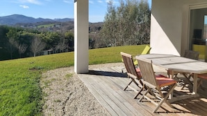 Alpendre coberto com mesa e cadeiras, com vista para uma área gramada e colinas ao longe.