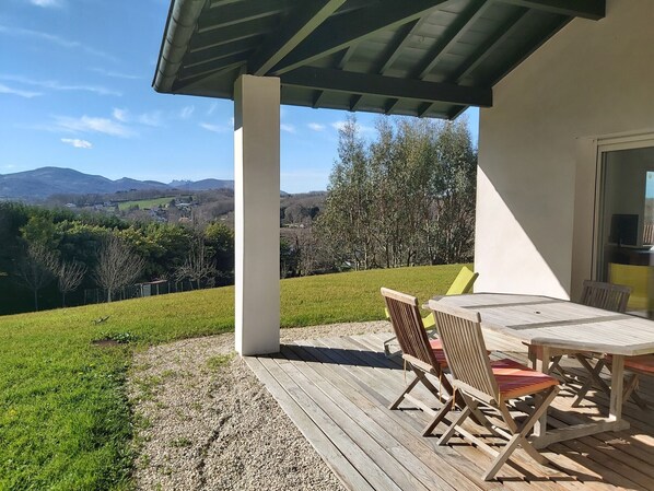Eine überdachte Terrasse mit Tisch und Stühlen, mit Blick auf eine grüne Wiese und ferne Hügel.