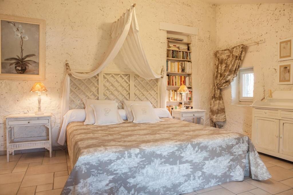 A bedroom with a canopy bed, a bedside table with a lamp, a bookshelf, and a window with curtains.