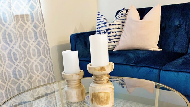 Ein runder Glastisch mit goldenen Metallbeinen, zwei Kerzen auf Holzständern, ein blaues Sofa mit gemusterten Kissen und ein Teppich mit Muster.