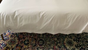 Ein ordentlich bezogenes Bett mit weißen Laken und zwei blauen Kissen, ein Nachttisch mit Lampe und ein gemustertes Teppich.