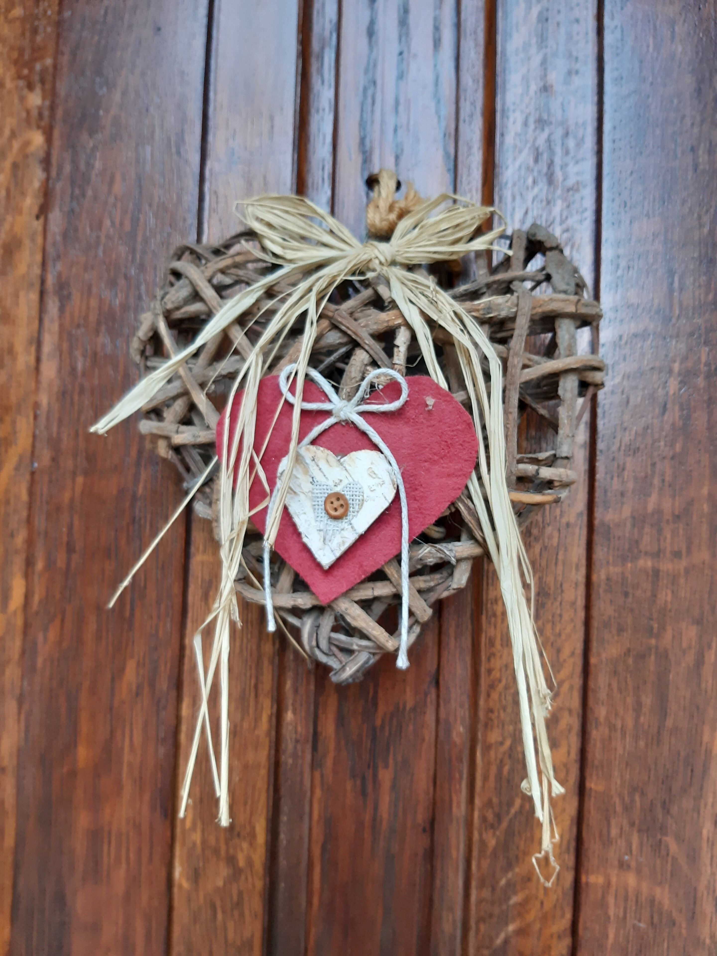 A rustic heart-shaped decoration made of twigs and straw, with a red felt heart and a white tag with a button.