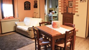A cozy living room with a white sofa, wooden dining table, and chairs. A radiator and a window with blinds are visible. - B&B Casa Mucci (Sestola)