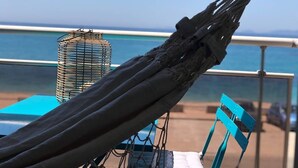 Eine Hängematte mit Metallgestell, ein blauer Stuhl und ein Tisch mit blauer Tischdecke, mit Blick auf einen Strand mit Geländer und dem Ozean im Hintergrund.