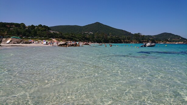 Ein Strand mit klarem Wasser, badenden Personen und Bergen im Hintergrund.