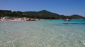 En strand med klart vatten, folk som badar och berg i bakgrunden.