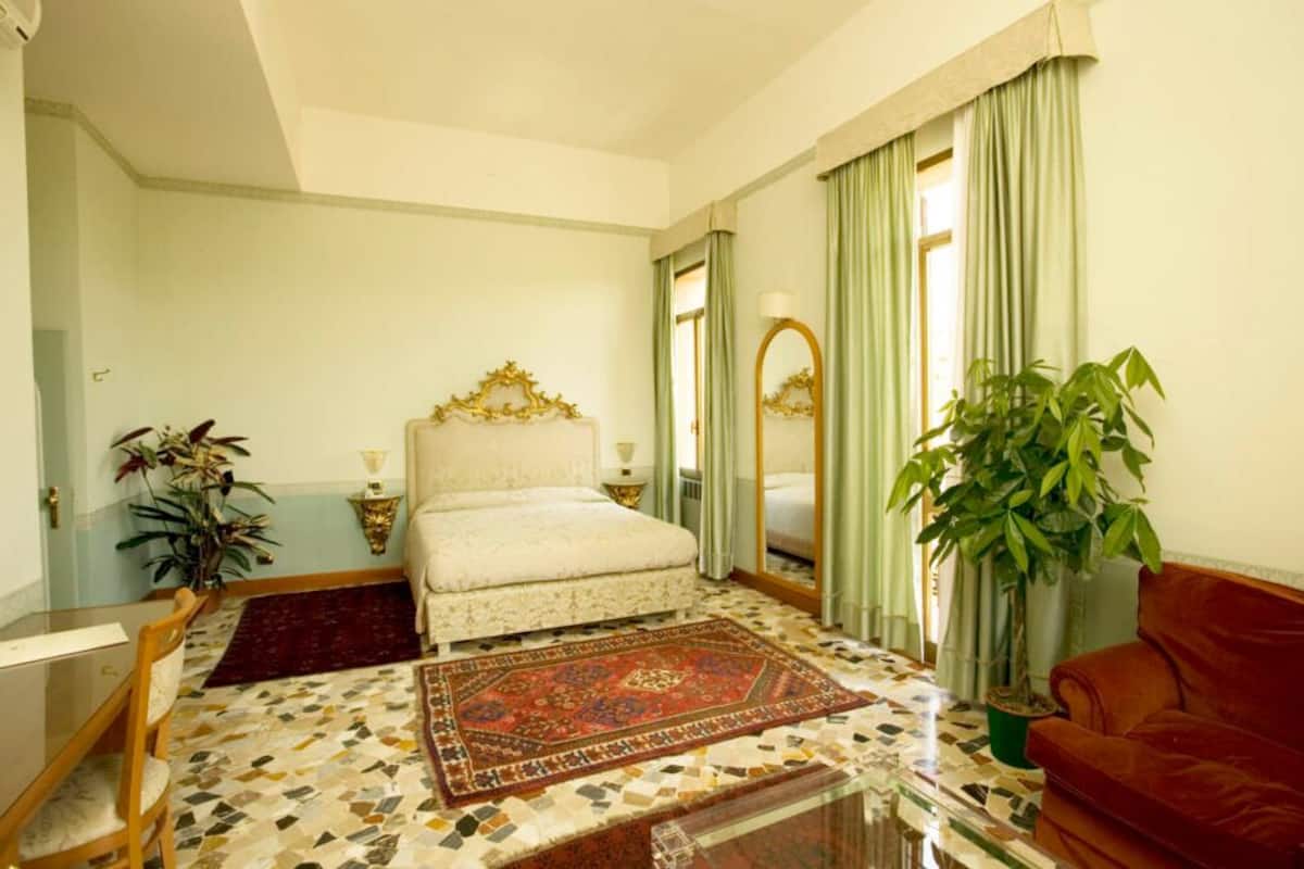 Ein Schlafzimmer mit Bett, einer Couch, einem großen Spiegel, einer Topfpflanze und einem gemusterten Teppich.