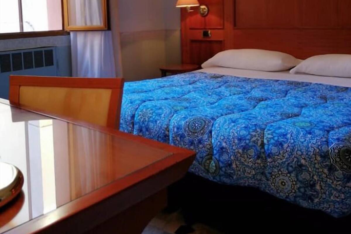 Ein Hotelzimmer mit Holz-Kopfteil, einem Bett mit blauen Bettbezug und Nachttisch mit Lampe sowie einem Fenster mit Vorhängen.