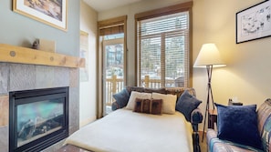A cozy room with a fireplace, a bed with patterned bedding, a window with blinds, and a lamp.