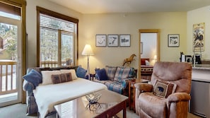 A cozy room with a bed, a sofa, a chair, and a dining table. There are framed pictures on the wall and a window with blinds.