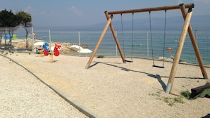 Una spiaggia con un'altalena in legno, un'area recintata con giochi per bambini e vista sul mare.