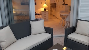 Un balcone con un divano e una poltrona, una cucina sullo sfondo e una porta scorrevole in vetro che dà su una camera da letto.