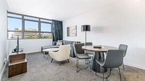 Een moderne woonkamer met een bank, twee fauteuils, een salontafel en een eettafel met stoelen. Er staat een tv op een dressoir, een vloerlamp en een schilderij aan de muur.