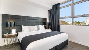 Een moderne hotelkamer met een groot bed, een hoofdeinde met een ingebouwde televisie, een nachtkastje met een lamp, en uitzicht op de stad door grote ramen.