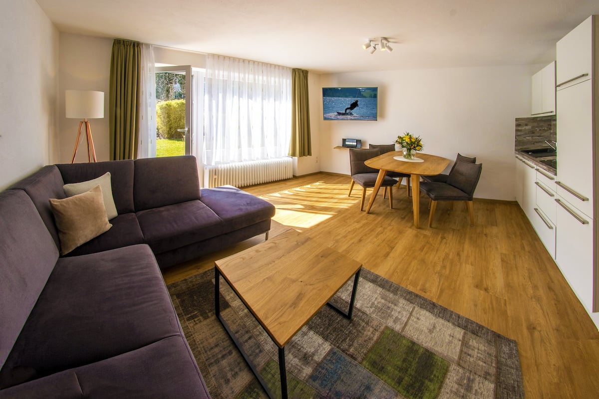 Un salon moderne avec un canapé, une table basse en bois et une table à manger avec des chaises. Il y a également un coin cuisine avec des placards et un réfrigérateur. Une fenêtre avec des rideaux laisse entrer la lumière naturelle dans la pièce.