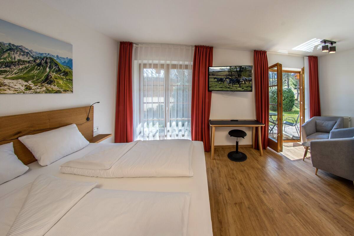 Une chambre d’hôtel avec un grand lit, un bureau, une chaise et un canapé. Il y a une fenêtre avec des rideaux, un tableau au mur et une vue sur l’extérieur.