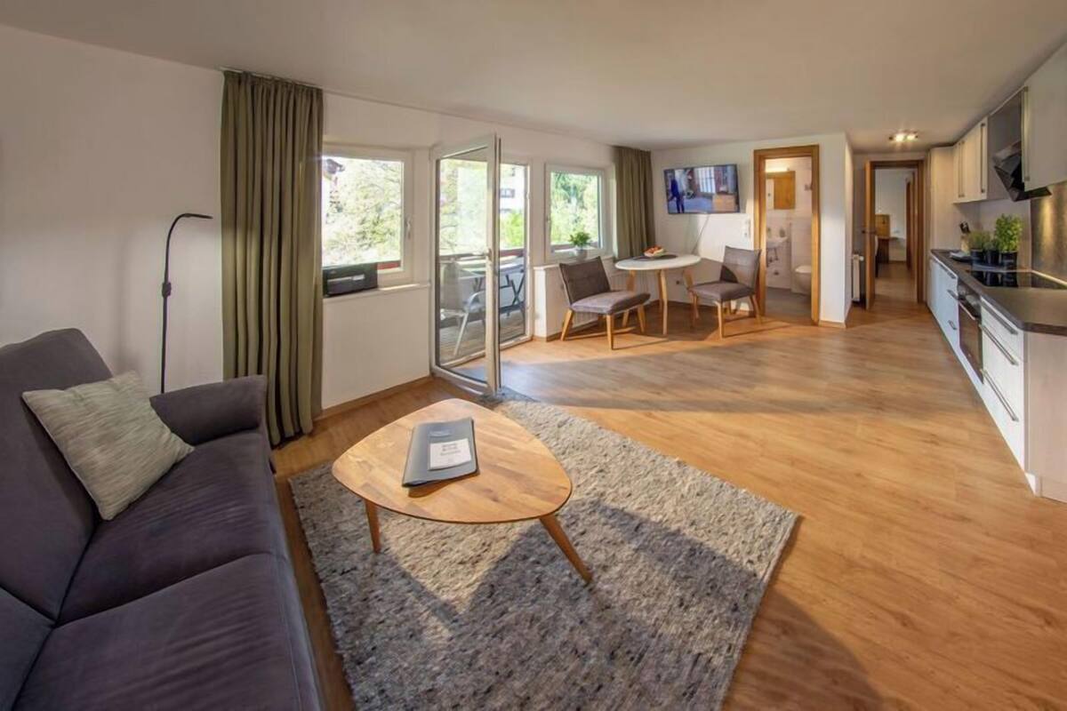 Un salon moderne avec un canapé foncé, une table basse en bois et un tapis gris. On y trouve également un coin repas avec une table et des chaises, ainsi qu’une cuisine équipée de placards et d’un téléviseur.